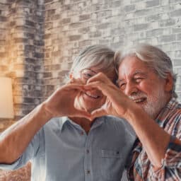 Close up of hands of two mature grandparents making a heart shape with their fingers together. Smiling and having fun in love people.