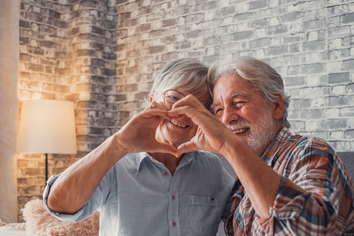 Close up of hands of two mature grandparents making a heart shape with their fingers together. Smiling and having fun in love people.